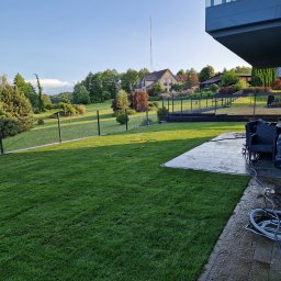 Nokop Andrzej Filipiak - Zadbany trawnik z systemem nawadniania i ogrodzeniem panelowym na tle domu z ogrodem. Widoczny fragment tarasu i nowoczesnej architektury budynku.