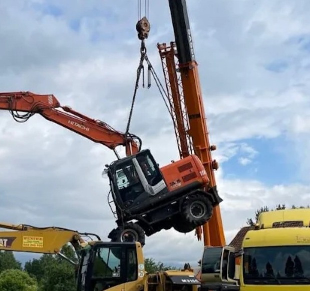 Pomarańczowa koparka Hitachi ZA 16 podnoszona przez dźwig na tle pochmurnego nieba, z widocznymi innymi maszynami budowlanymi w tle.