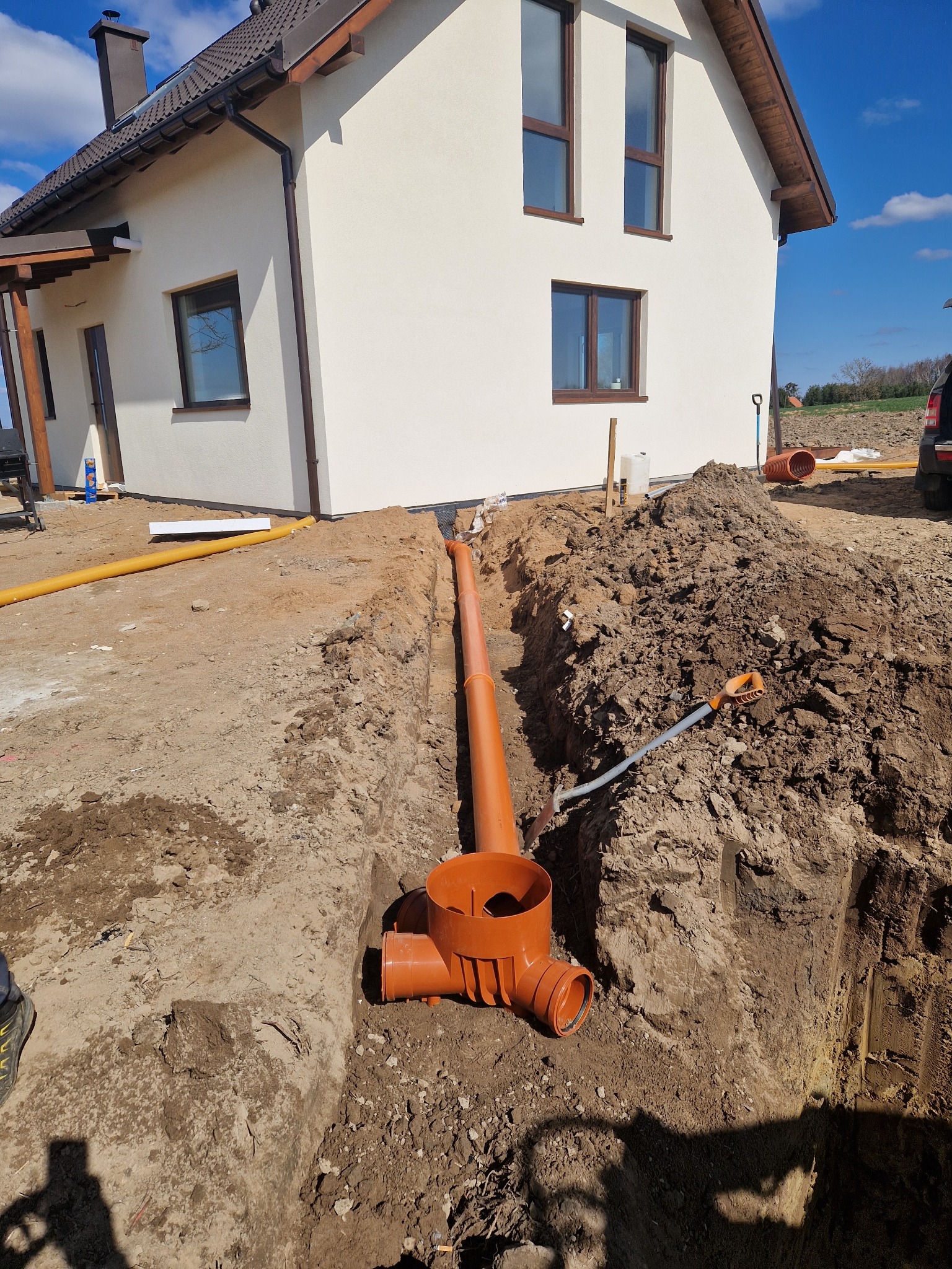 Pomarańczowe rury kanalizacyjne ułożone w wykopie ziemnym obok jasnego domu z brązowymi oknami, widoczne narzędzia i żółta rura drenażowa. Słoneczny dzień.