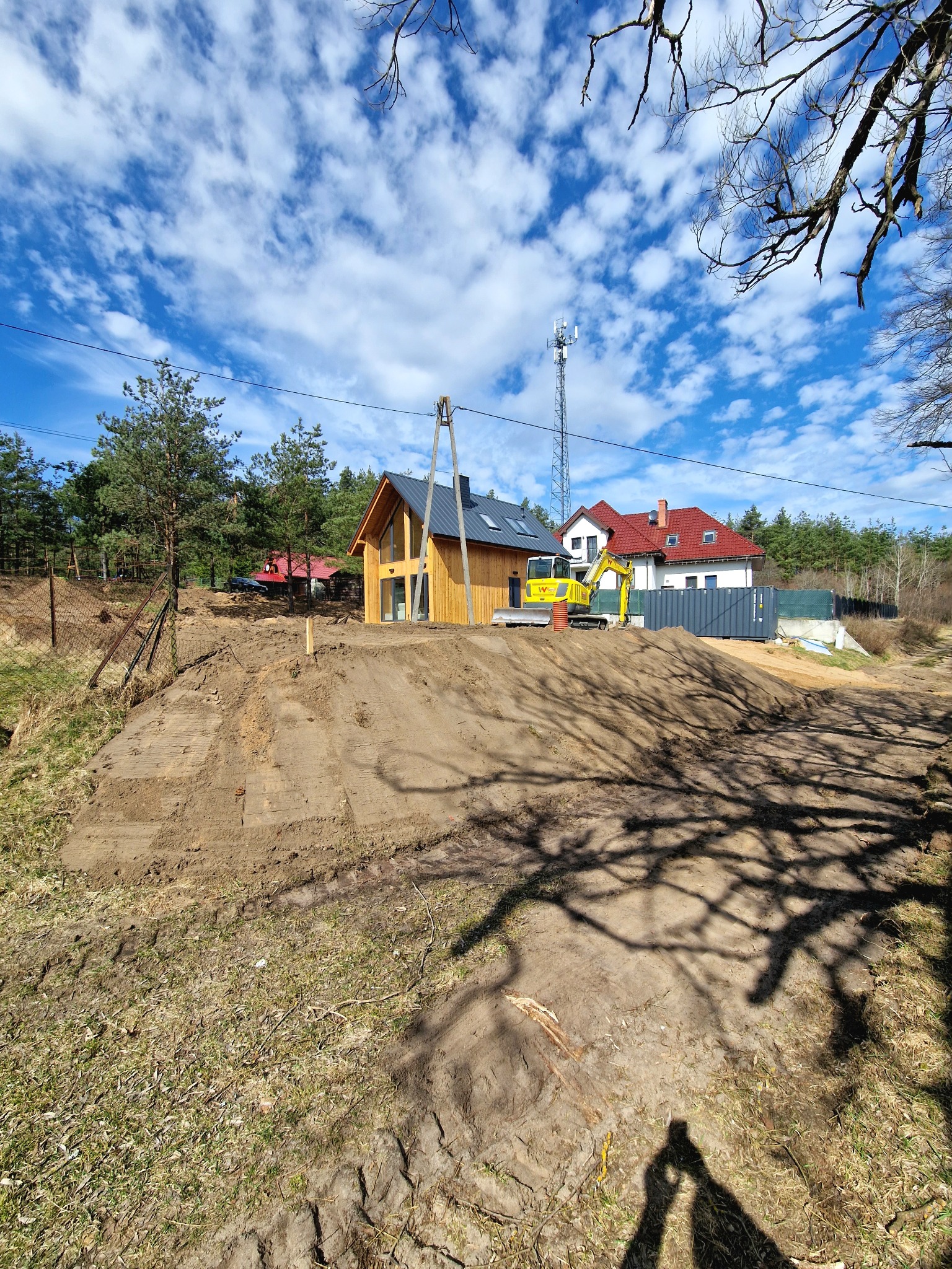 Żółta koparka na wzgórzu z przygotowanym terenem pod budowę domu. W tle domy, drzewa i błękitne niebo z chmurami. Cień drzewa w dolnej części kadru.