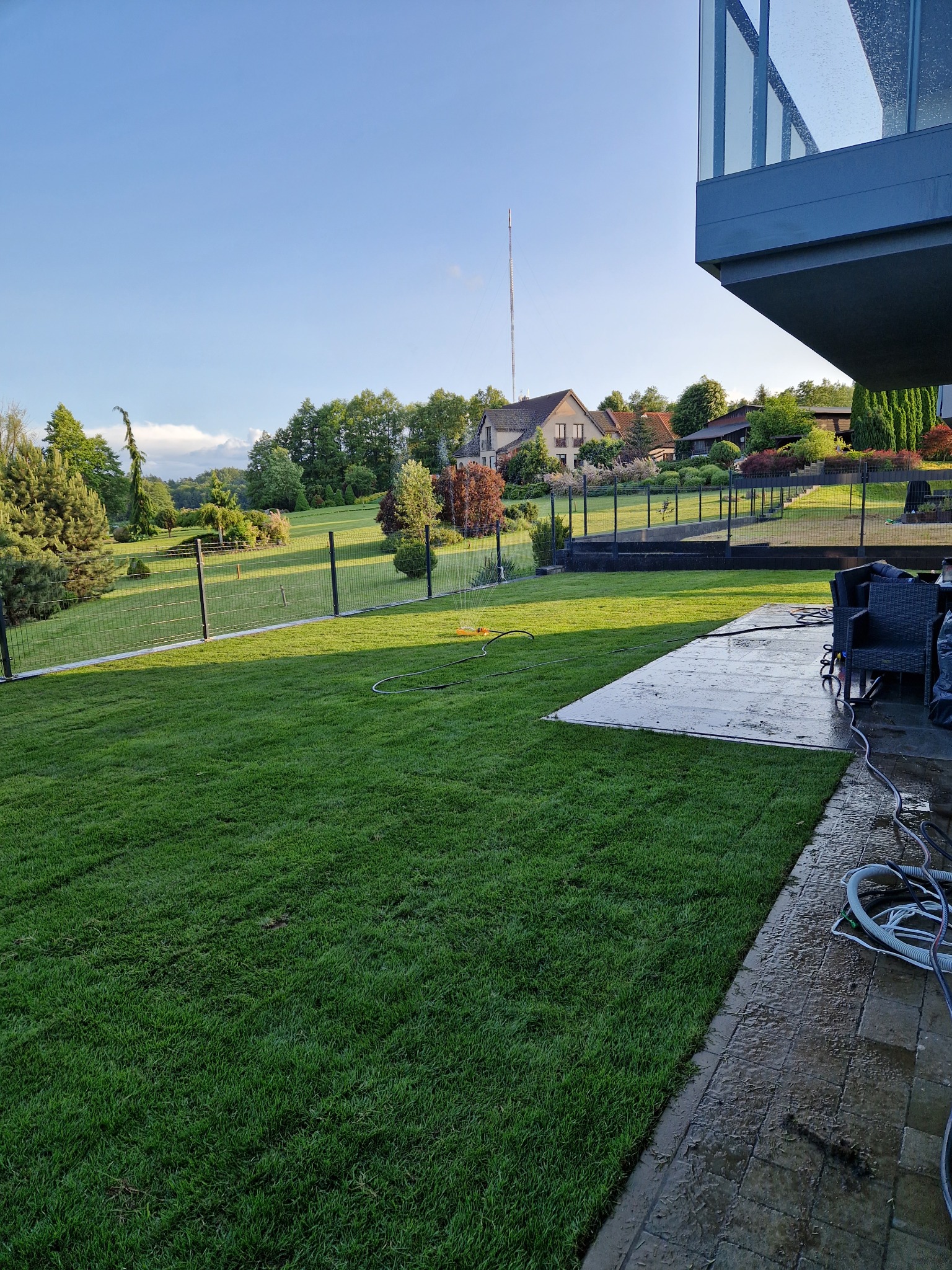Zadbany trawnik z systemem nawadniania i ogrodzeniem panelowym na tle domu z ogrodem. Widoczny fragment tarasu i nowoczesnej architektury budynku.