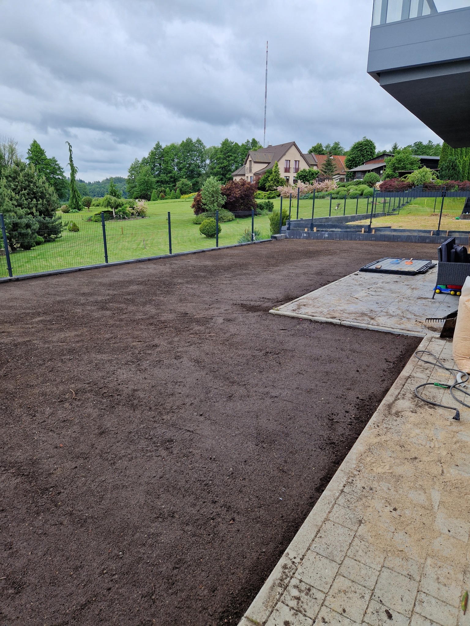 Przygotowany teren pod trawnik z widoczną siatką ogrodzeniową i fragmentem domu w tle. Wyrównana ziemia, gotowa do siewu trawy. Widoczne narzędzia ogrodnicze.