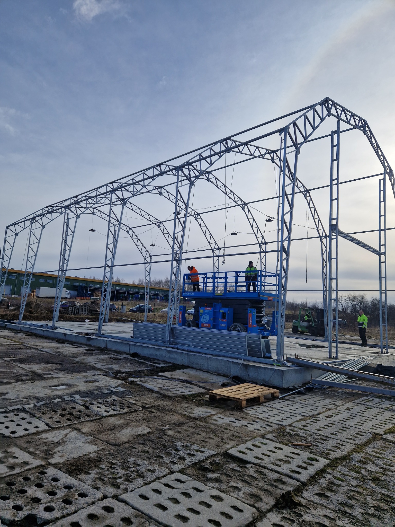 Montaż konstrukcji stalowej hali w Olsztynie. Widoczni pracownicy na podnośniku koszowym, montujący szkielet hali. Prace budowlane w toku, widoczne elementy konstrukcyjne.