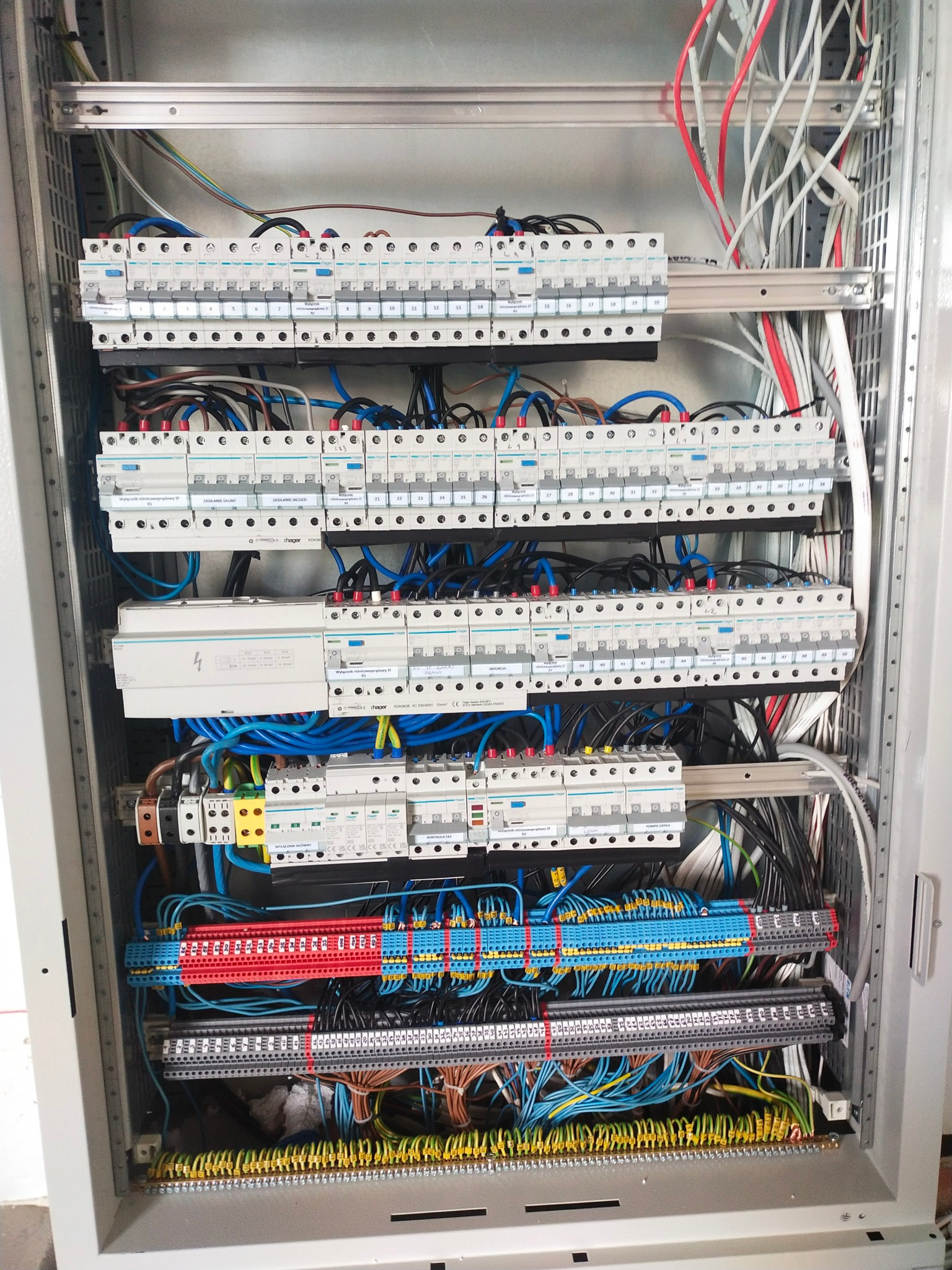Rozdzielnica elektryczna z licznymi zabezpieczeniami, okablowaniem w różnych kolorach i listwami zaciskowymi, zamontowana w metalowej szafie. Widok z bliska.