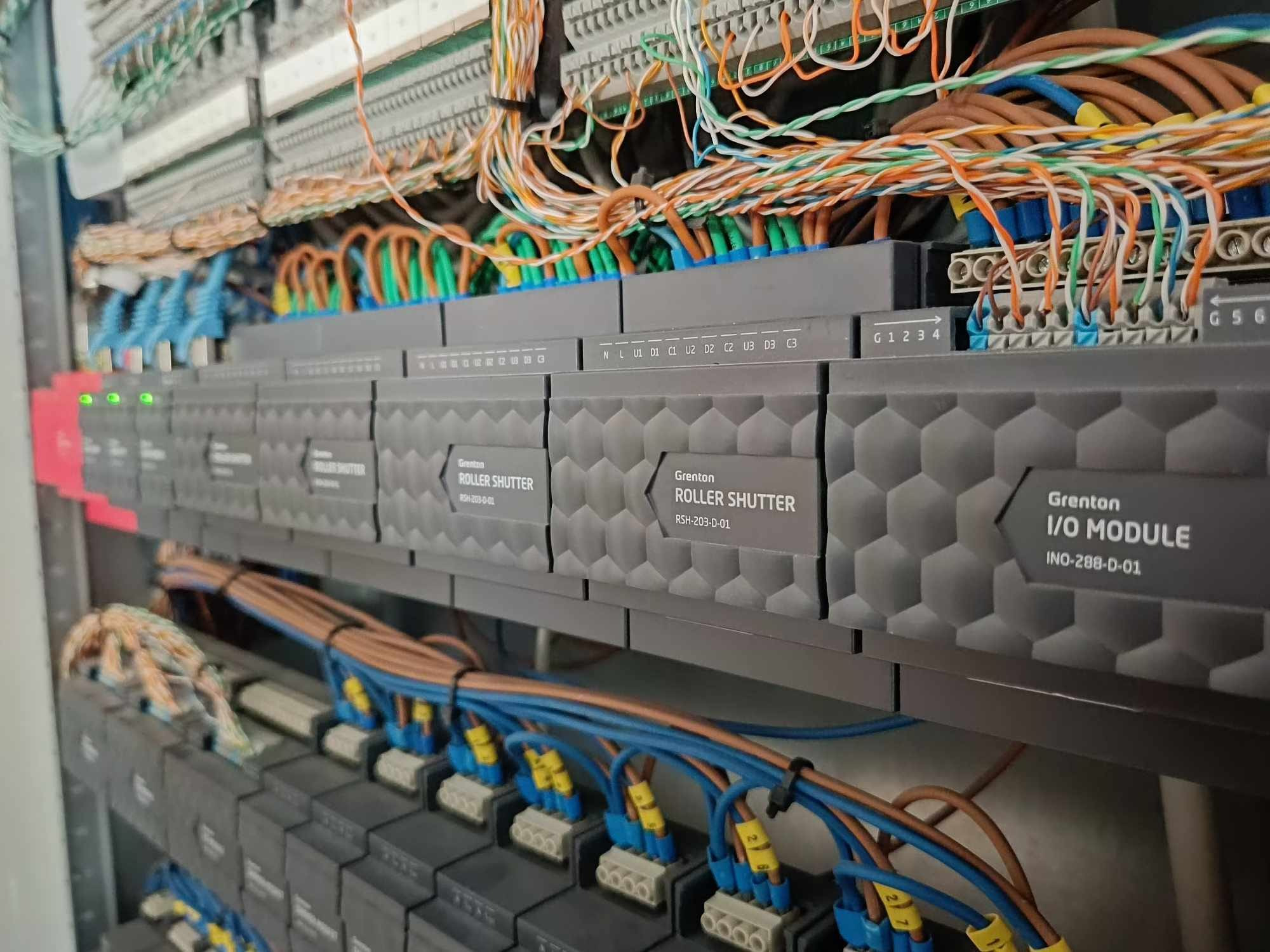 Szare moduły sterujące roletami Grenton Roller Shutter RSH-203-D-01 i moduł I/O Module INO-288-D-01, z podłączonymi przewodami w różnych kolorach, w szafie sterowniczej.