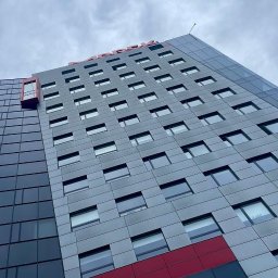 City View Nieruchomości - Agnieszka Sadowska - Wysoki budynek biurowy z szarą fasadą i licznymi oknami, widziany z dołu pod kątem. Na szczycie widoczny czerwony napis. Część fasady jest szklana.