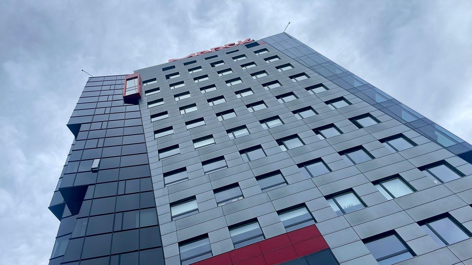 Wysoki budynek biurowy z szarą fasadą i licznymi oknami, widziany z dołu pod kątem. Na szczycie widoczny czerwony napis. Część fasady jest szklana.