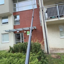 Pure Glass Maciej Malinowski - Czyszczenie okien na wysokości za pomocą teleskopowego kija z dyszą, widok z dołu na budynek z balkonami i zielenią. Usługa sprzątania.