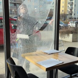 Pure Glass Maciej Malinowski - Mycie okna w lokalu gastronomicznym. Widoczne smugi po myciu, budynki w tle. Na pierwszym planie stolik z krzesłami. Osoba myjąca okno częściowo widoczna.
