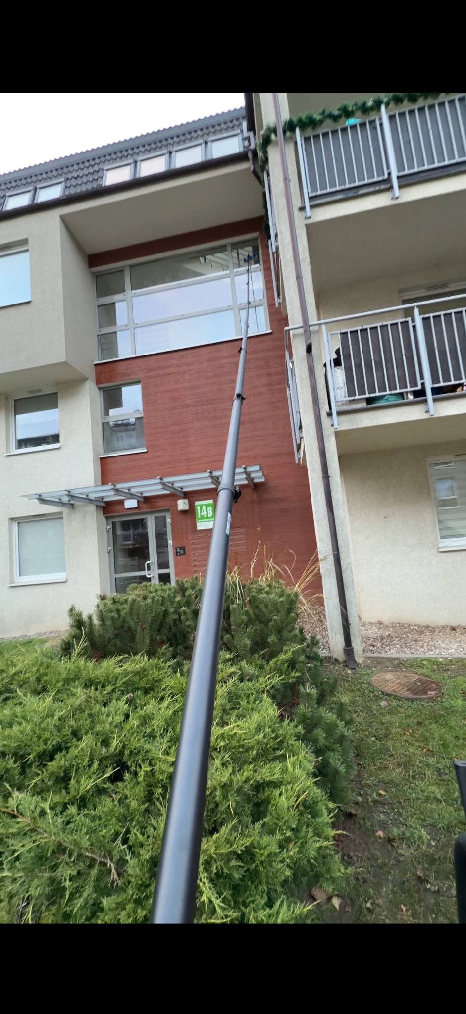 Czyszczenie okien na wysokości za pomocą teleskopowego kija z dyszą, widok z dołu na budynek z balkonami i zielenią. Usługa sprzątania.