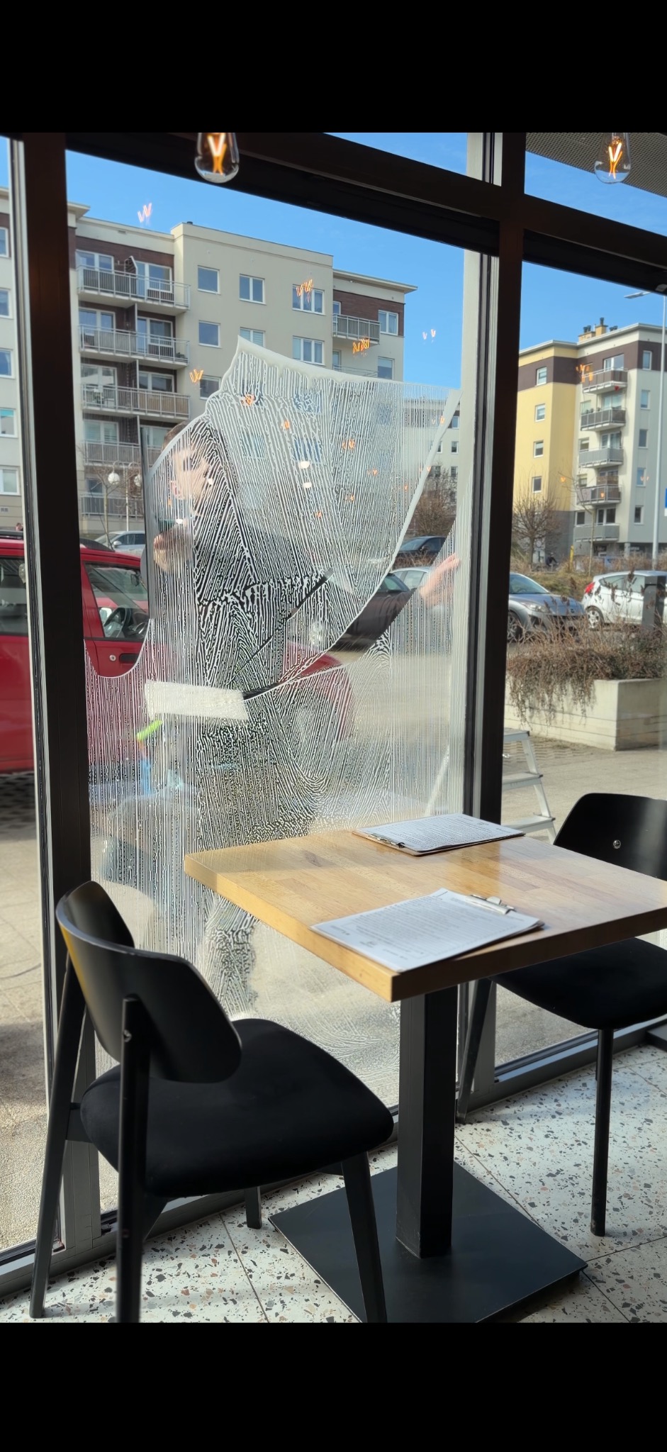 Mycie okna w lokalu gastronomicznym. Widoczne smugi po myciu, budynki w tle. Na pierwszym planie stolik z krzesłami. Osoba myjąca okno częściowo widoczna.