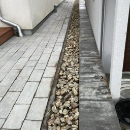 PATRYK MARZEC - Długi, wąski pas kamieni oddziela chodnik z szarych płyt od ściany budynku. Widok z góry, perspektywa zbieżna w oddali. Minimalistyczny design ogrodu.
