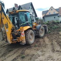 FHU WETERAN Damian Kozina - Żółta koparko-ładowarka JCB na nierównym, błotnistym terenie z pryzmą ziemi w tle, domy w oddali. Widoczne ślady opon w błocie.