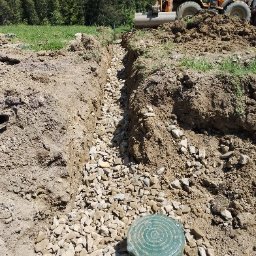 FHU WETERAN Damian Kozina - Wąski wykop wypełniony kamieniami, z zieloną studzienką na pierwszym planie i koparko-ładowarką w tle, na tle zieleni i drzew. Prace ziemne w terenie pagórkowatym.