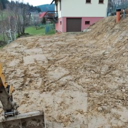 FHU WETERAN Damian Kozina - Żółta koparko-ładowarka częściowo widoczna, przygotowuje teren pod budowę domu na wzgórzu. W tle budynek z garażem i drzewa.