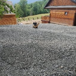 FHU WETERAN Damian Kozina - Utwardzona, kamienista powierzchnia z zagęszczarką w tle, otoczona drewnianymi budynkami i zielonymi wzgórzami. Widok na krajobraz górski.