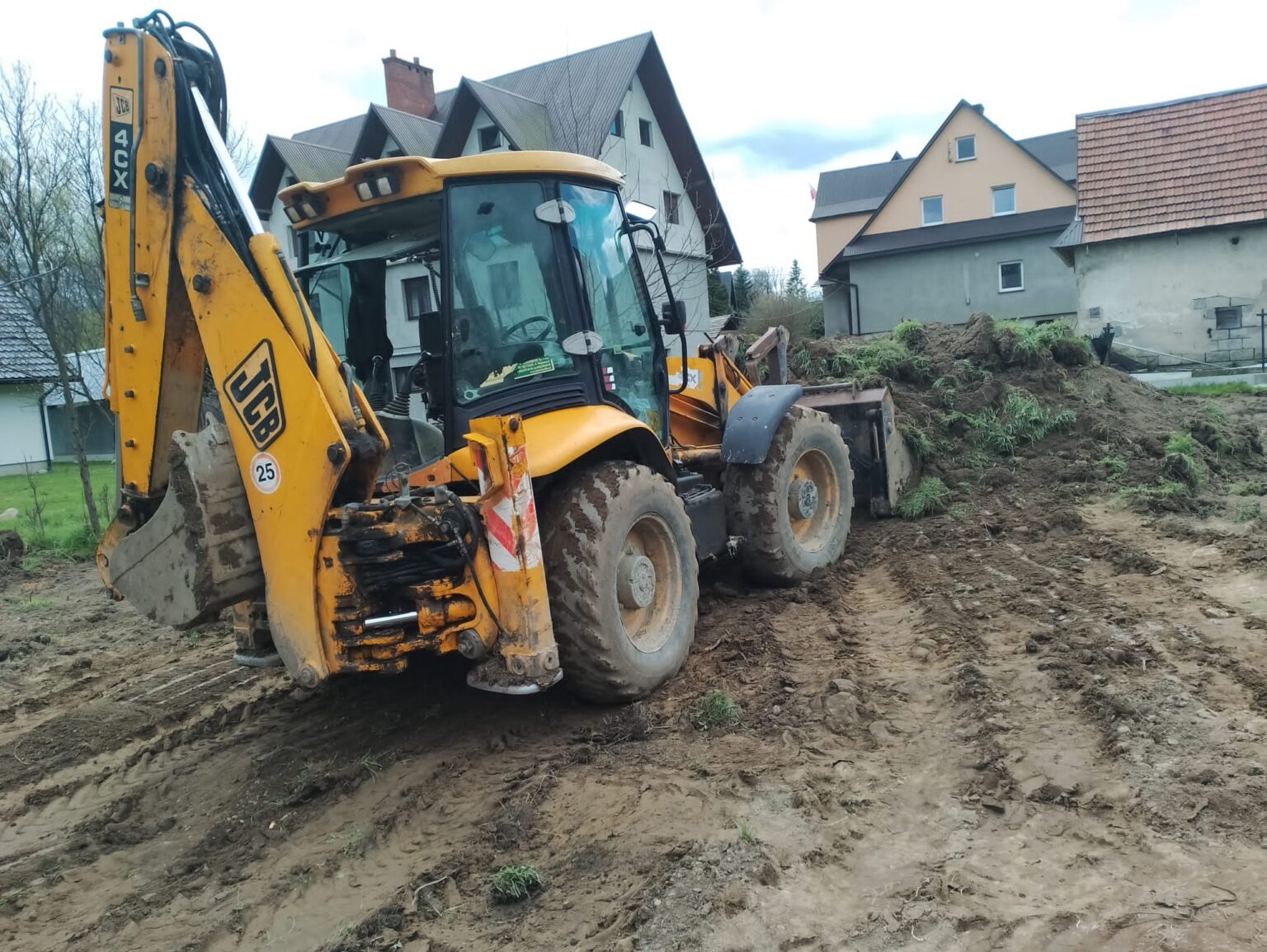 Żółta koparko-ładowarka JCB na nierównym, błotnistym terenie z pryzmą ziemi w tle, domy w oddali. Widoczne ślady opon w błocie.