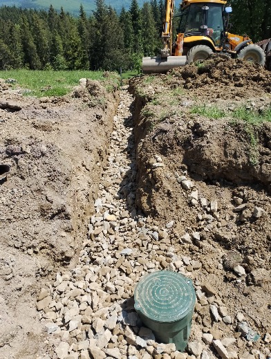 Wąski wykop wypełniony kamieniami, z zieloną studzienką na pierwszym planie i koparko-ładowarką w tle, na tle zieleni i drzew. Prace ziemne w terenie pagórkowatym.