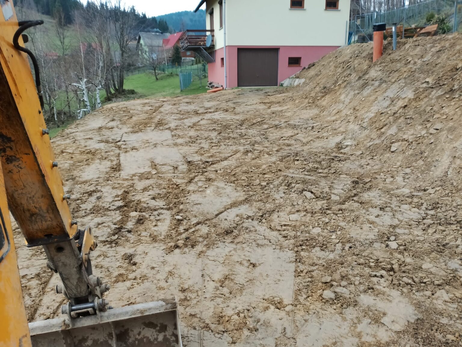 Żółta koparko-ładowarka częściowo widoczna, przygotowuje teren pod budowę domu na wzgórzu. W tle budynek z garażem i drzewa.