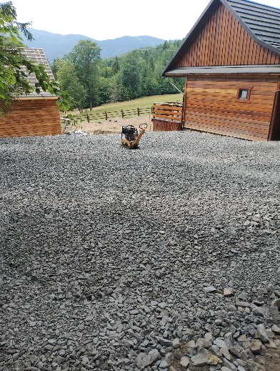 Utwardzona, kamienista powierzchnia z zagęszczarką w tle, otoczona drewnianymi budynkami i zielonymi wzgórzami. Widok na krajobraz górski.