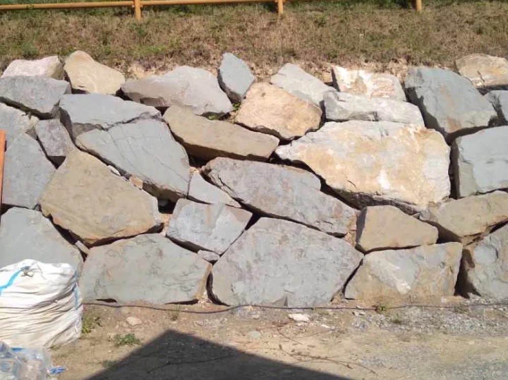 Ułożony mur z nieregularnych, szarych i beżowych kamieni, zabezpieczający skarpę. W tle trawa i drewniane barierki. U dołu widoczny worek z materiałem.