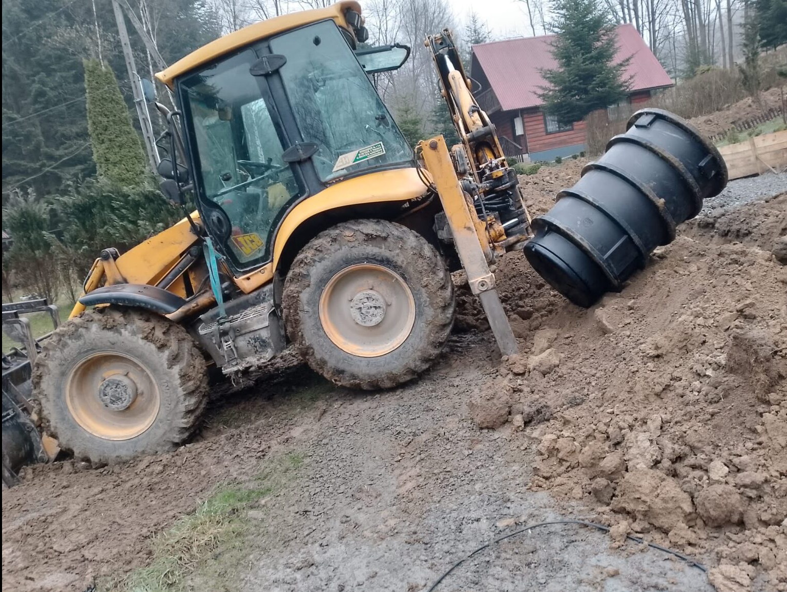 Żółta koparko-ładowarka umieszcza czarny zbiornik w wykopie na tle domu z czerwonym dachem i drzew. Błotniste podłoże, pochmurne niebo.