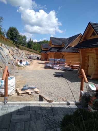 Budowa domów z bali w górskim krajobrazie. Widoczne materiały budowlane, utwardzony teren i kamienny mur oporowy. Słoneczny dzień, błękitne niebo z chmurami.