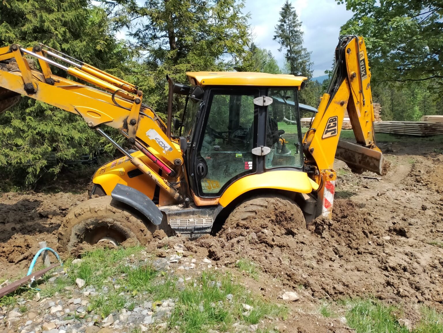 Żółta koparko-ładowarka JCB ugrzęzła w błocie na terenie budowy. Widoczne ślady opon i stos drewna w tle. Ujęcie z bliska, podkreślające trudne warunki pracy.