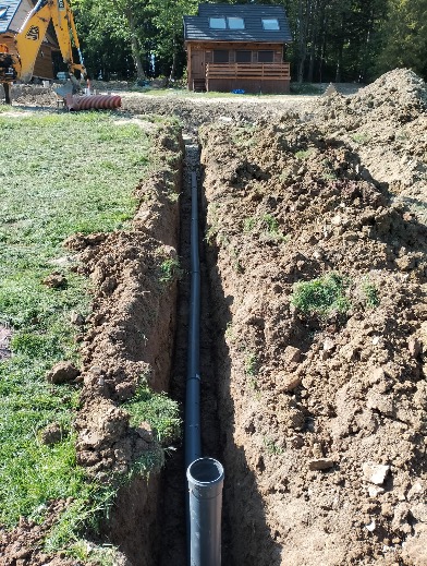 Wąski wykop z ułożoną rurą kanalizacyjną na tle domku i koparki. Widok z góry na dół, oświetlenie dzienne, ziemia piaszczysta.