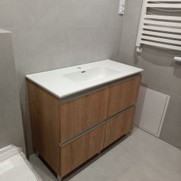 Gajda Meble na wymiar - Nowoczesna szafka łazienkowa z umywalką nablatową, fronty w odcieniach drewna, szare płytki, minimalistyczny grzejnik drabinkowy. Ujęcie z niskiego kąta.