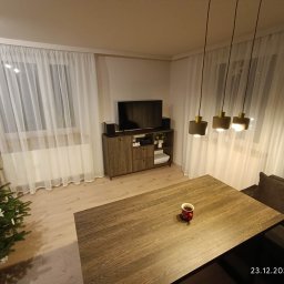 Gajda Meble na wymiar - Nowoczesny salon z drewnianą komodą pod telewizorem, zasłonami i choinką. Na stole stoi kubek. Ciepłe oświetlenie wnętrza.