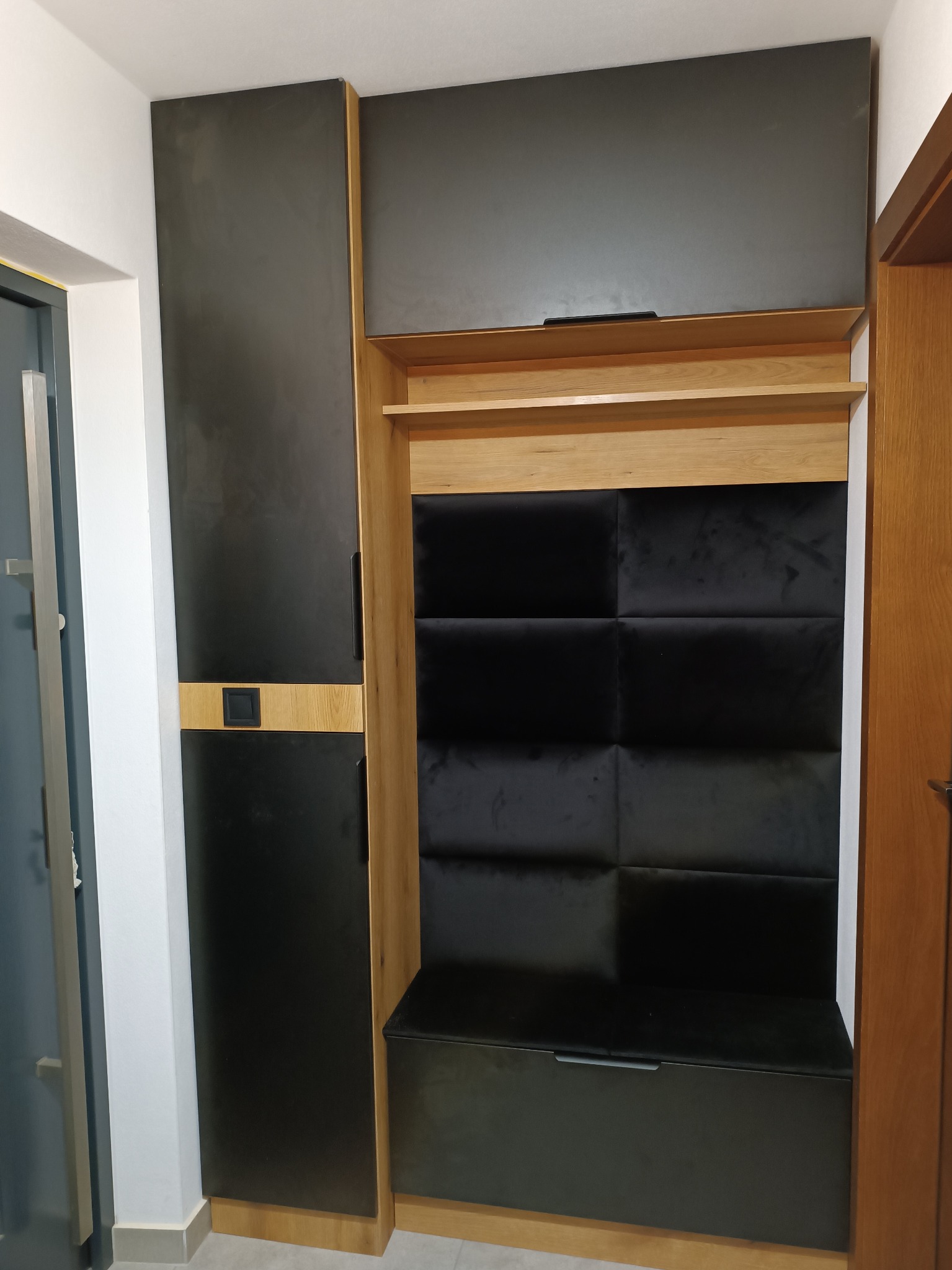 Nowoczesna szafa wnękowa z czarnymi frontami i drewnianymi akcentami, z siedziskiem i tapicerowanym panelem. Minimalistyczny design, oświetlenie punktowe.