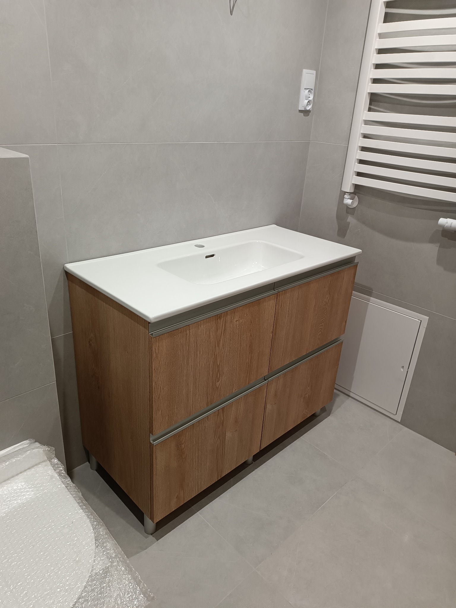 Nowoczesna szafka łazienkowa z umywalką nablatową, fronty w odcieniach drewna, szare płytki, minimalistyczny grzejnik drabinkowy. Ujęcie z niskiego kąta.