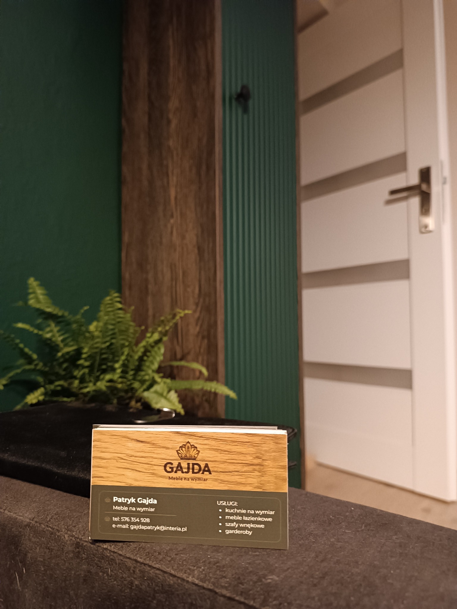 Wizytówka firmy meblarskiej 'GAJDA Meble na wymiar' na tle zielonej ściany z drewnianymi elementami i rośliną doniczkową. Widoczna oferta: kuchnie, szafy wnękowe.