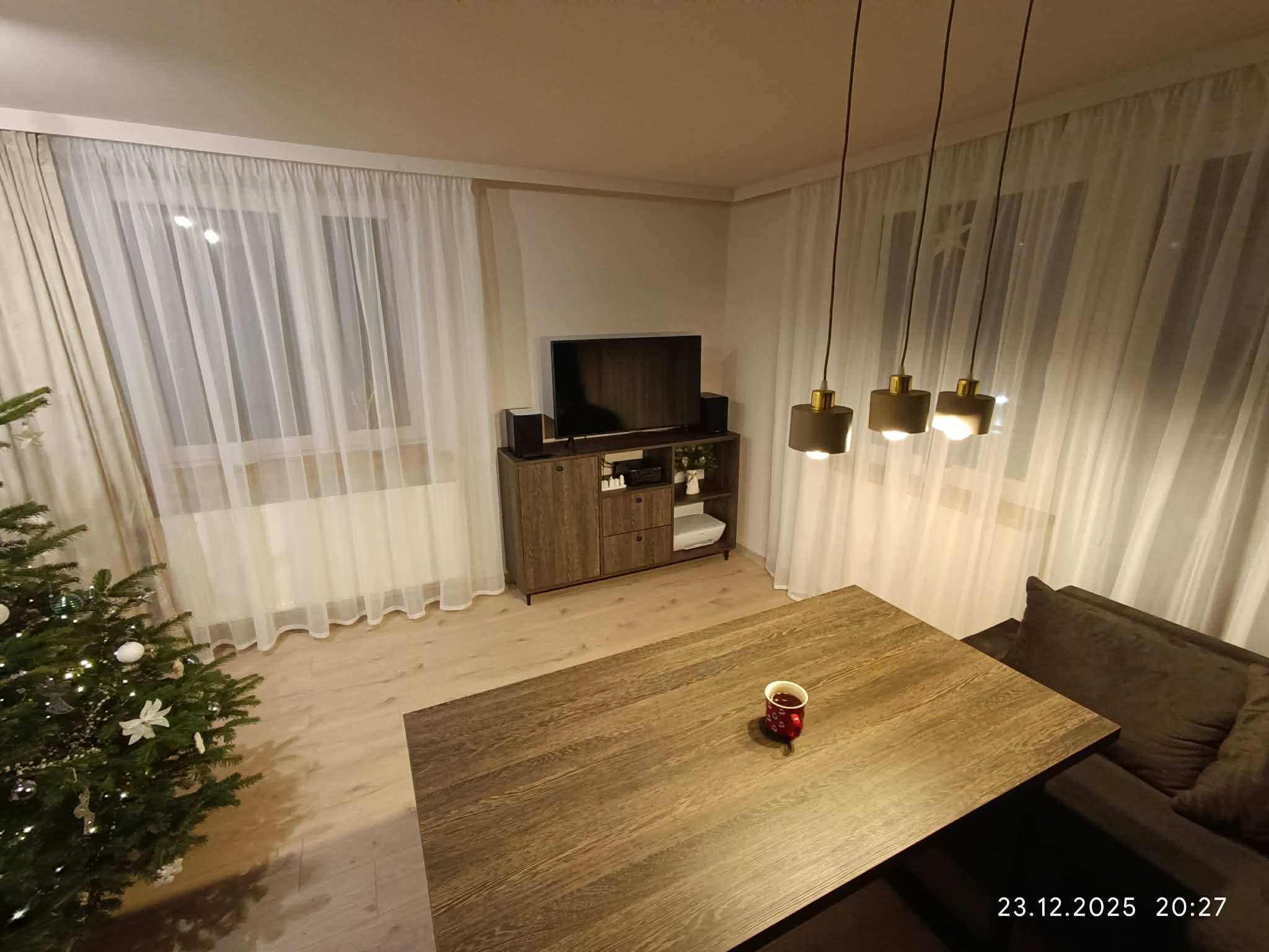 Nowoczesny salon z drewnianą komodą pod telewizorem, zasłonami i choinką. Na stole stoi kubek. Ciepłe oświetlenie wnętrza.