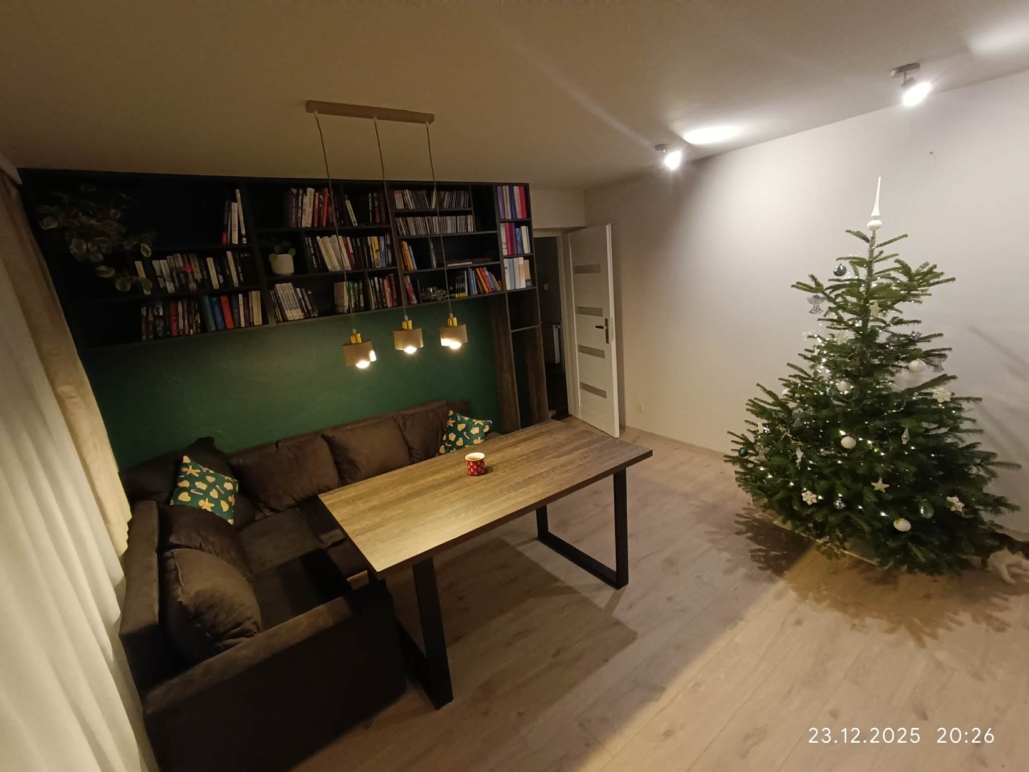 Nowoczesny salon z narożną sofą, drewnianym stołem na metalowych nogach i regałem na książki w zabudowie. Świąteczny klimat podkreśla choinka z ozdobami.