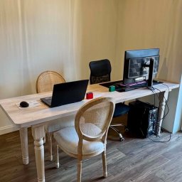 Verano Insurance - Jasne biuro z drewnianym biurkiem, laptopem, monitorem i krzesłami. Widok na zewnątrz przez okno. Minimalistyczny design, domowe biuro.
