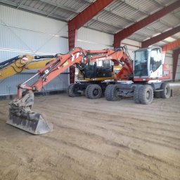 Dremex - Dwie koparki kołowe Terex Atlas w hali w Elblągu. Pomarańczowa i żółta maszyna, widoczne ślady pracy na piasku. Konstrukcja stalowa hali w tle.