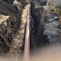 Dremex - Brązowa rura ułożona w wykopie ziemnym, widoczny pracownik w tle. Wykop z czarną ziemią i piaskiem, narzędzia pracy obok. Instalacja podziemna w trakcie realizacji.