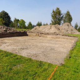 Dremex - Wyrównany teren pod budowę z wykopem i hałdami ziemi. W tle zieleń drzew i częściowo widoczny biały samochód dostawczy. Pomarańczowa linia wyznacza granicę.