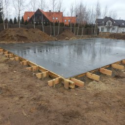 Dremex - Wylana płyta fundamentowa z drewnianym szalunkiem na działce budowlanej. Widoczne pryzmy ziemi i zabudowa jednorodzinna w tle. Szara, pochmurna pogoda.