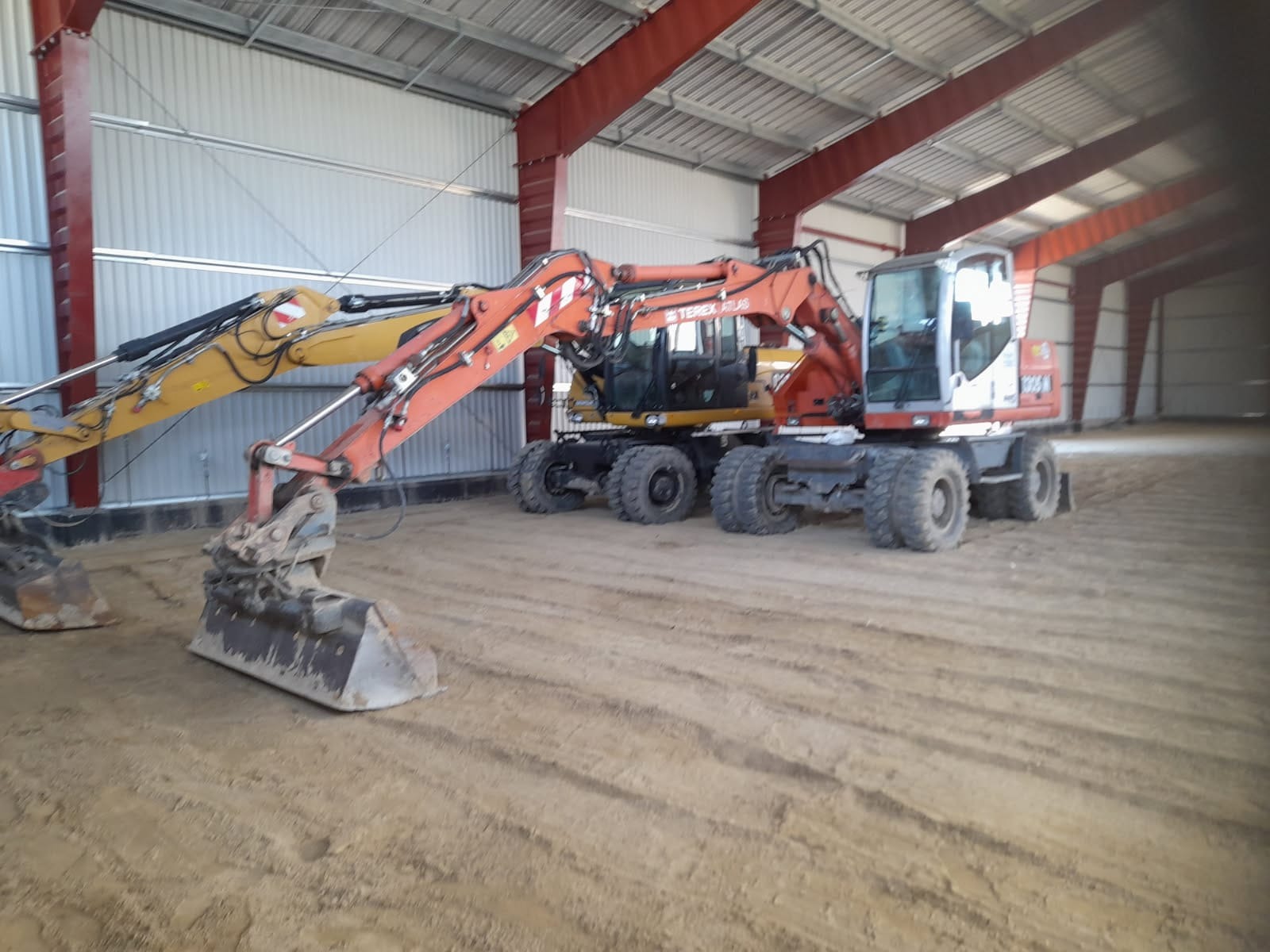 Dwie koparki kołowe Terex Atlas w hali w Elblągu. Pomarańczowa i żółta maszyna, widoczne ślady pracy na piasku. Konstrukcja stalowa hali w tle.