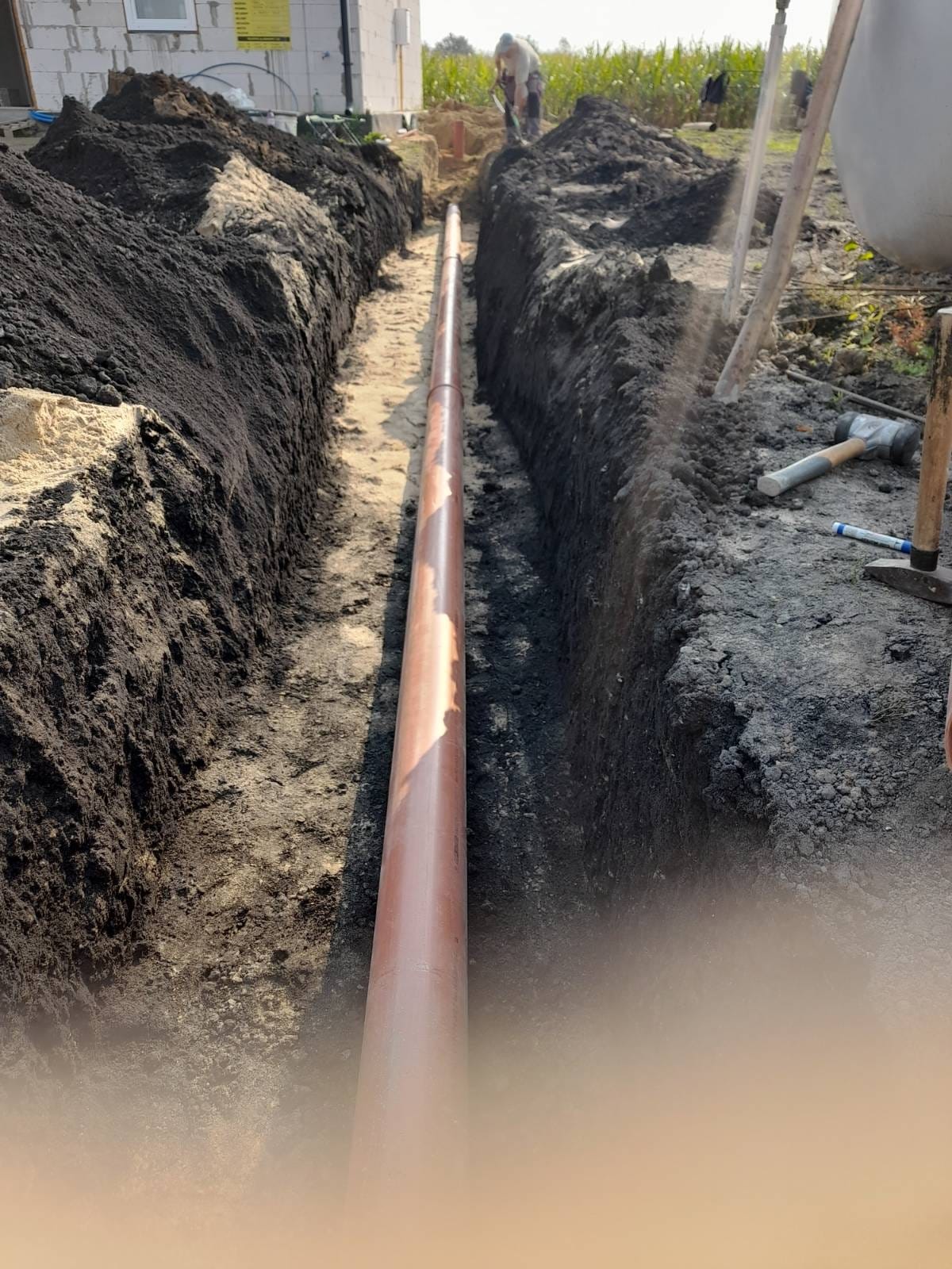 Brązowa rura ułożona w wykopie ziemnym, widoczny pracownik w tle. Wykop z czarną ziemią i piaskiem, narzędzia pracy obok. Instalacja podziemna w trakcie realizacji.