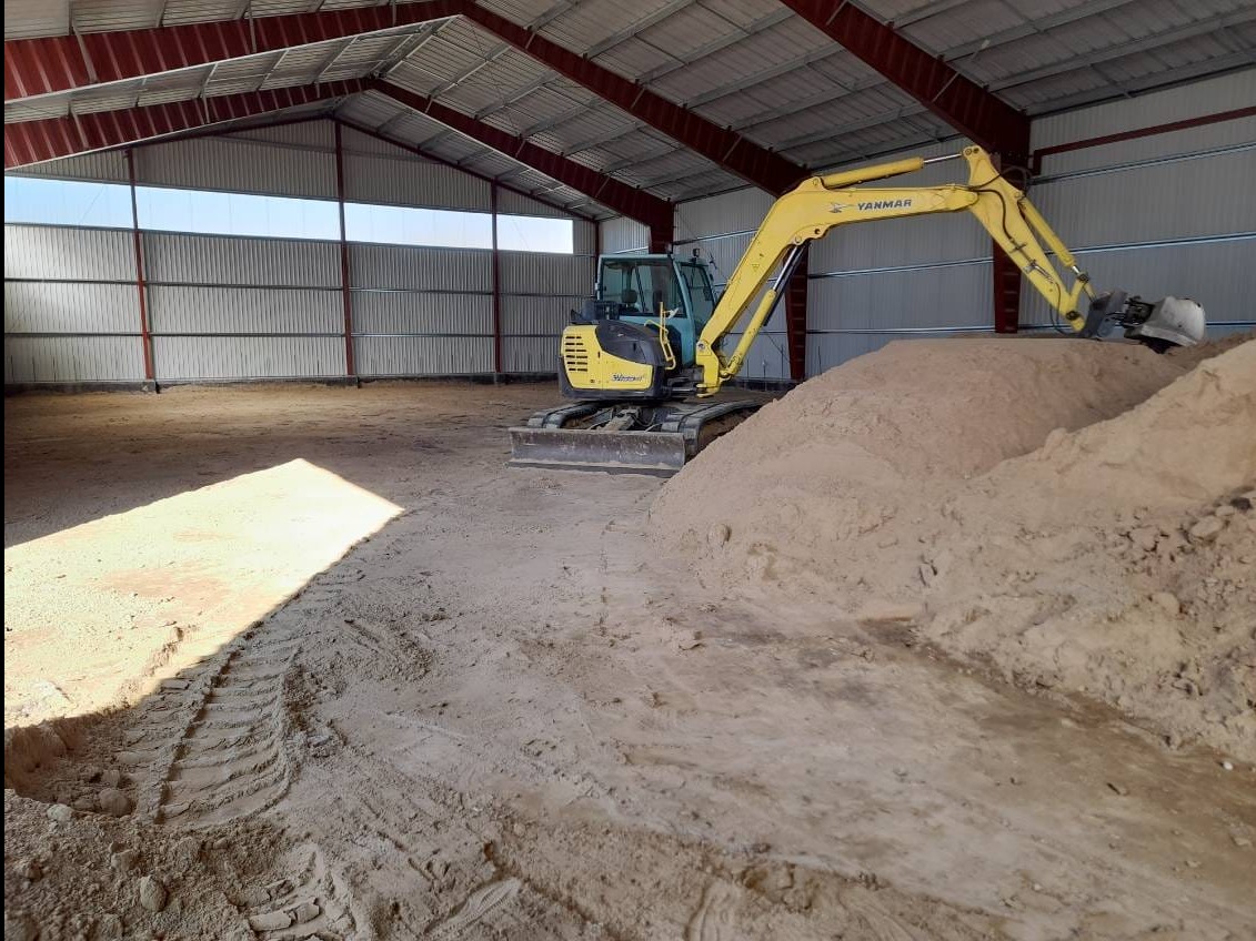 Żółta mini koparka Yanmar w hali z metalową konstrukcją, obok sterta piasku. Widoczne ślady opon na piasku. Hala oświetlona naturalnym światłem.