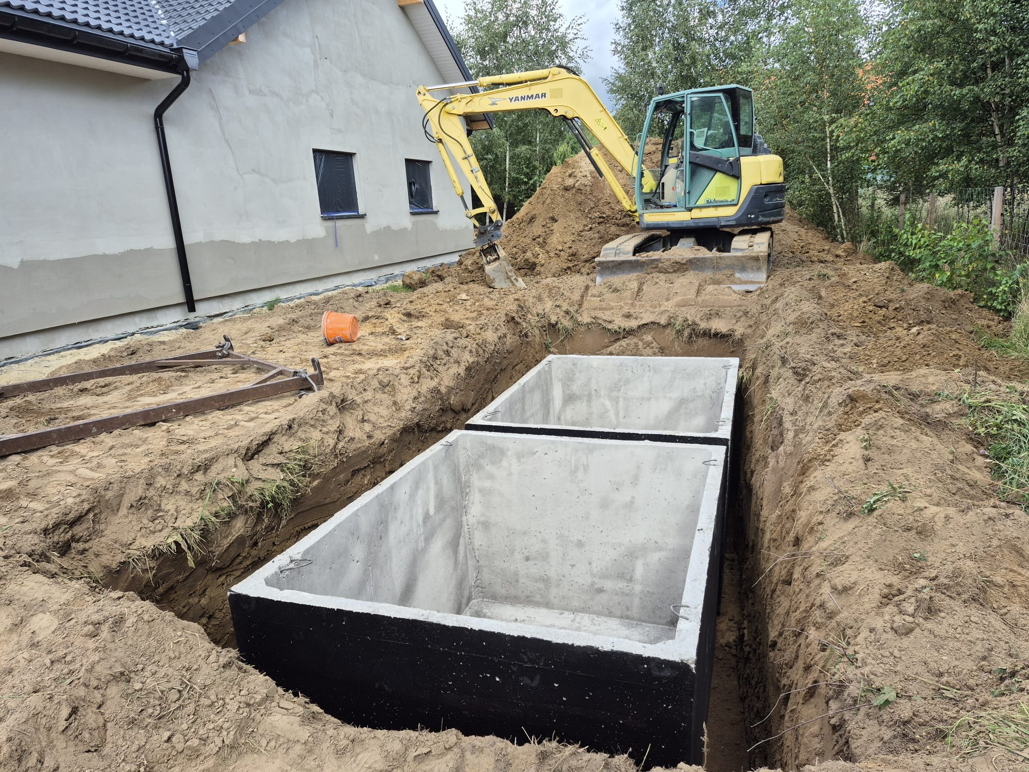 Dwa betonowe zbiorniki w wykopie, przygotowane do instalacji szamba. Żółta koparka Yanmar w tle, obok budynku mieszkalnego. Widok z góry na dół.