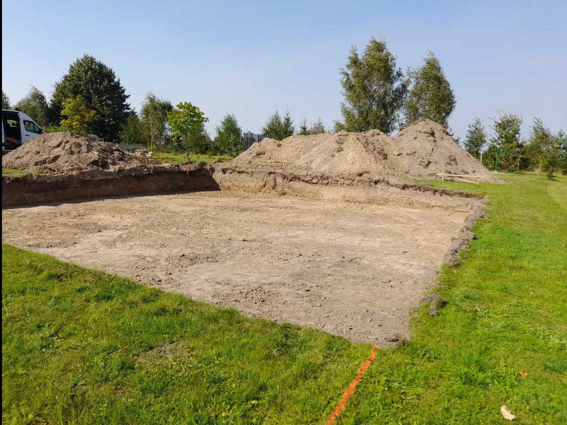 Wyrównany teren pod budowę z wykopem i hałdami ziemi. W tle zieleń drzew i częściowo widoczny biały samochód dostawczy. Pomarańczowa linia wyznacza granicę.
