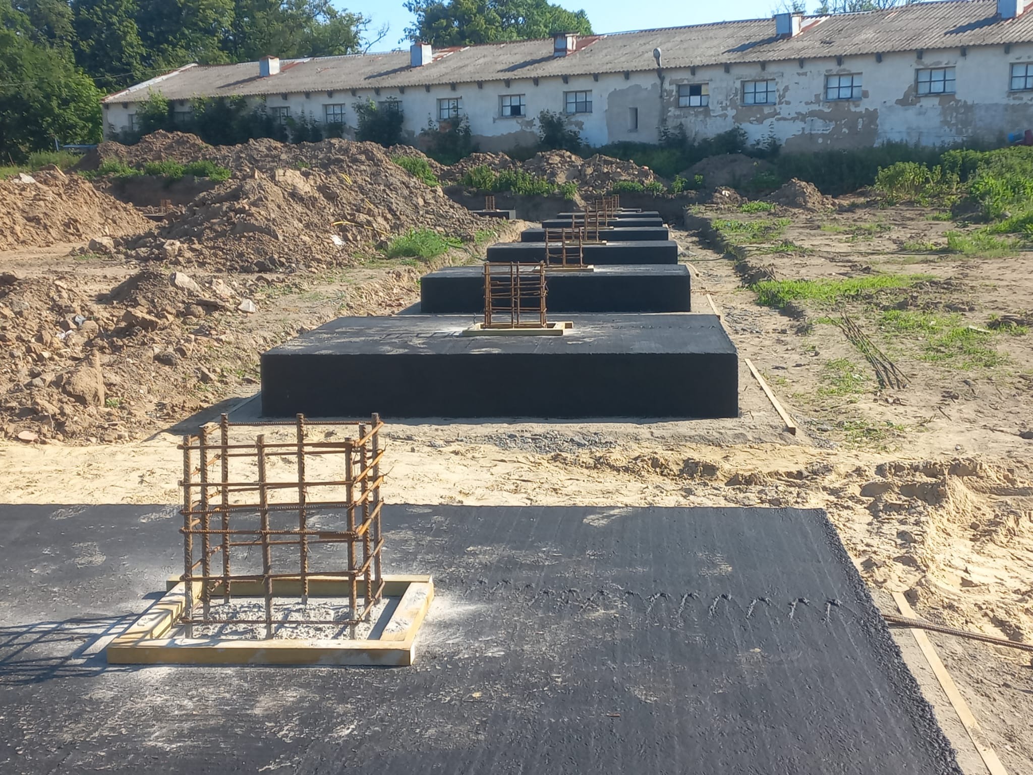 Ułożone, czarne płyty fundamentowe z wystającymi zbrojeniami na piaszczystym terenie budowy. W tle budynek z białej cegły i hałda ziemi.