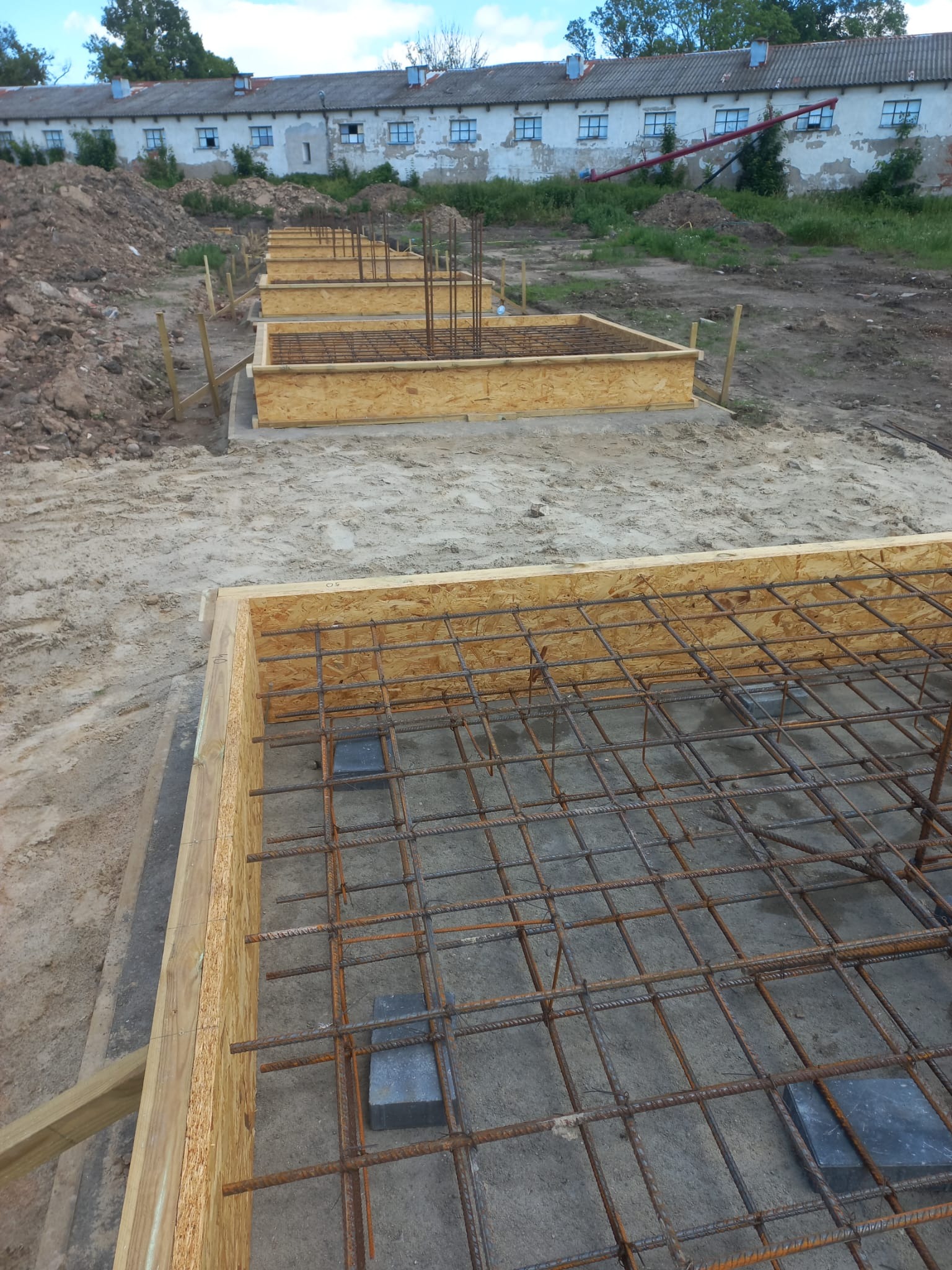 Przygotowane szalunki i zbrojenie pod fundamenty na piaszczystym podłożu. Widoczne drewniane formy i stalowe pręty zbrojeniowe, w tle budynek gospodarczy i hałda ziemi.