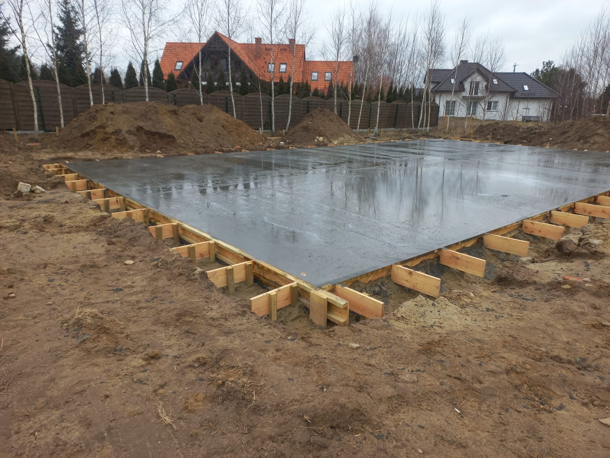 Wylana płyta fundamentowa z drewnianym szalunkiem na działce budowlanej. Widoczne pryzmy ziemi i zabudowa jednorodzinna w tle. Szara, pochmurna pogoda.