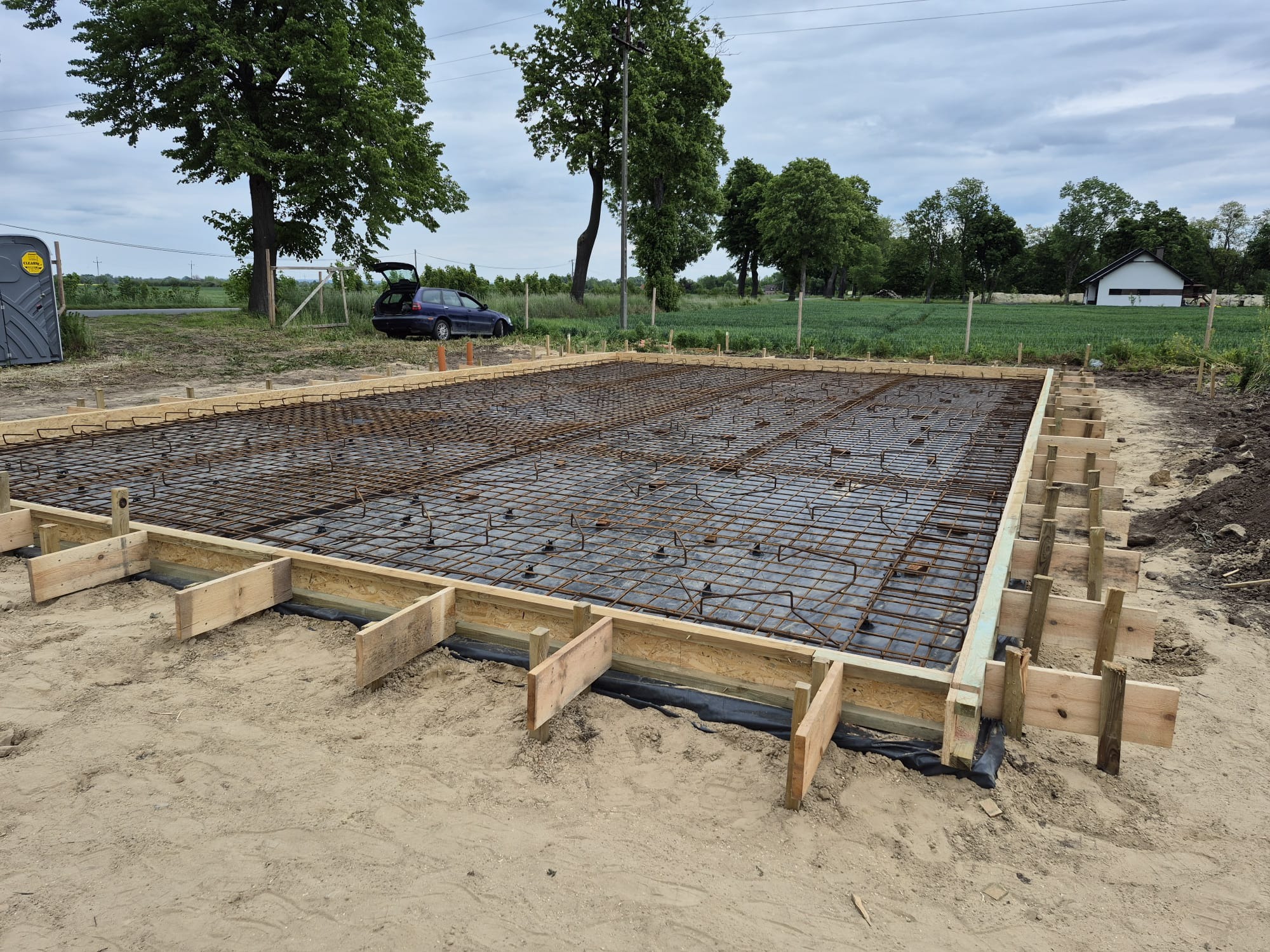 Zbrojenie fundamentu z drewnianym szalunkiem na piaszczystym podłożu, widok z góry. W tle drzewa, samochód i budynek.